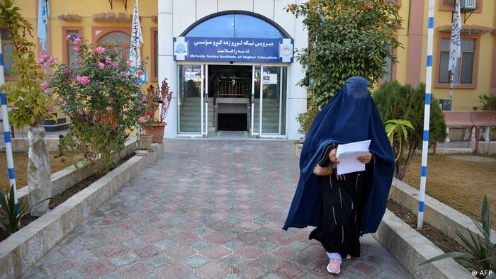 Das Bild zeigt den Eingang zu einer Universität in der afghanischen Provinz Kandahar, flankiert von Büschen und Rosensträuchern. Eine in eine dunkelblaue Burka gehüllte Frau entfernt sich vom Eingang, in der Hand hält sie einen Stapel Papiere.