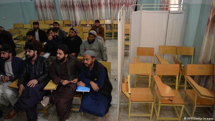 Links im Bild eine Gruppe männlicher Studierender, die offenbar an einem Seminar teilnimmt. Rechts, abgeteilt durch einen Vorhang, stehen sechs leere Stühle.