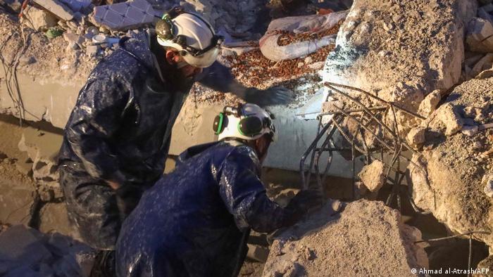 Two men in protective clothing and white helmets peer into a gap between building rubble