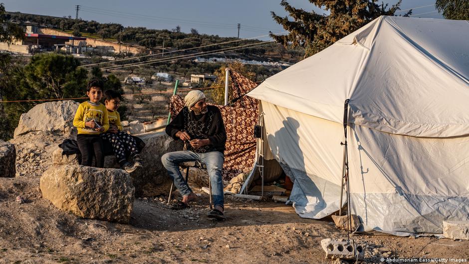 رجل وطفلان يجلسون خارج خيمة مؤقتة في مخيم في إدلب - سوريا. A man and two children sit outside a makeshift tent in a camp in Idlib (image: Abdulmonam Eassa/Getty Images)