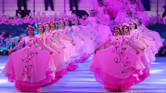 Two rows of women in pink ballgowns stand holding their skirts out on a stage.