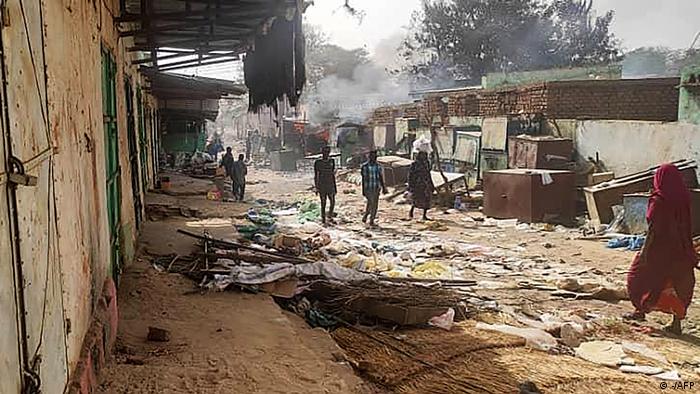 People walking along a dirt road with rubbish and things lying around, smoke rising in the background.