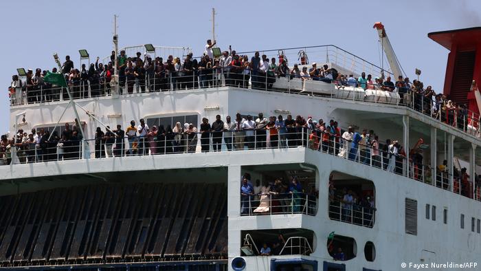 Hundreds of people stand at the railing on the decks of a ferry above the raised loading ramp.