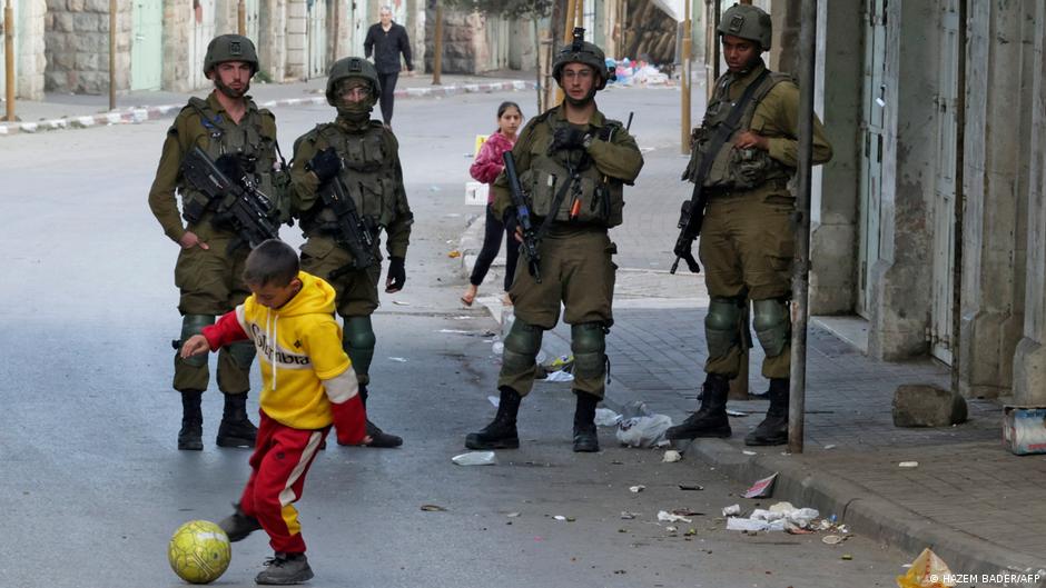 A Palestinian child plays football in the street in Hebron in front of four Israeli soldiers