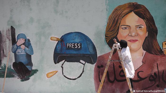 Eine Person malt an einem überlebensgroßen Wandgemälde, das Akle im Porträt und in Schutzausrüstung zeigt, außerdem einen Presse-Helm durchbohrt von einer Gewehrkugel