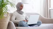 A smiling Muslim woman wearing a hijab sits and looks at her laptop